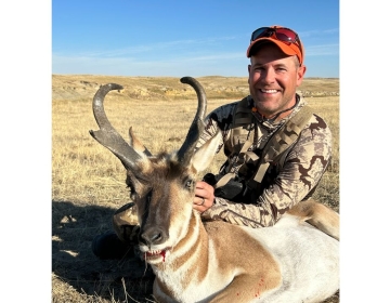 A hunter wearing a camouflage jacket and orange hat smiles while holding an impressive antelope trophy, captured during an SNS Outfitter & Guides hunt.